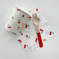 A paper cup, napkin and fork. The cherry paper party cups and napkins feature a red, green and white cherry print. The wooden fork is dipped in red.