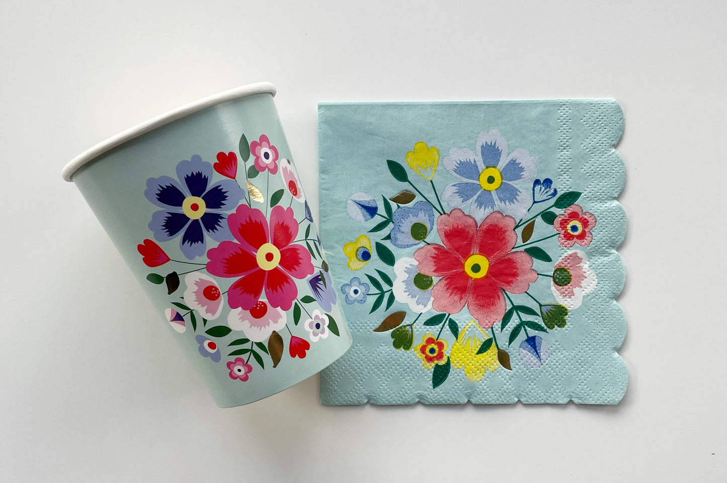 The Blossom Party Kit paper party napkins and paper cups. The flower/floral pattern includes green, red and blue colours. The napkins have scalloped edges.