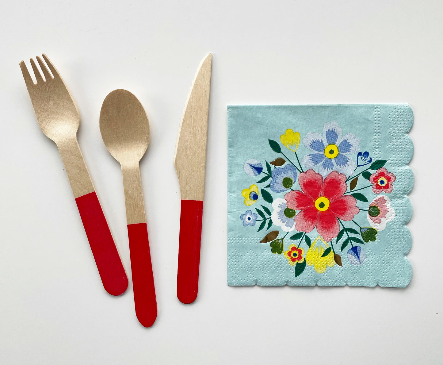 The Blossom Party Kit's napkins and utensils. The paper party napkins have a flower/floral pattern including green, red and blue colours. The napkins have scalloped edges. The Blossom party kit's red dipped wooden utensils, including a fork, spoon and knife.