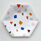 The large Terrazzo paper party plates are in the shape of a hexagon. The terrazzo pattern includes blue, gold, orange and green colours.  