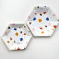 The large and small Terrazzo paper party plates are in the shape of a hexagon. The terrazzo pattern includes blue, gold, orange and green colours.  