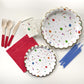 The complete Celebration Party Kit including large paper party plates, small paper party plates, paper cups, red paper napkins, red dipped wooden utensils and tall blue birthday candles. The celebration pattern features green, orange and blue balloons, ice cream cones, party hats, party garlands, birthday gifts and donuts. The party plates feature gold scalloped edges.