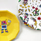 Close up of the large and small robot paper party plates. They are in the shape of a dodecagon. The robot pattern includes planets, robots, spaceships and clouds in green, blue, red, yellow and pink colours.  