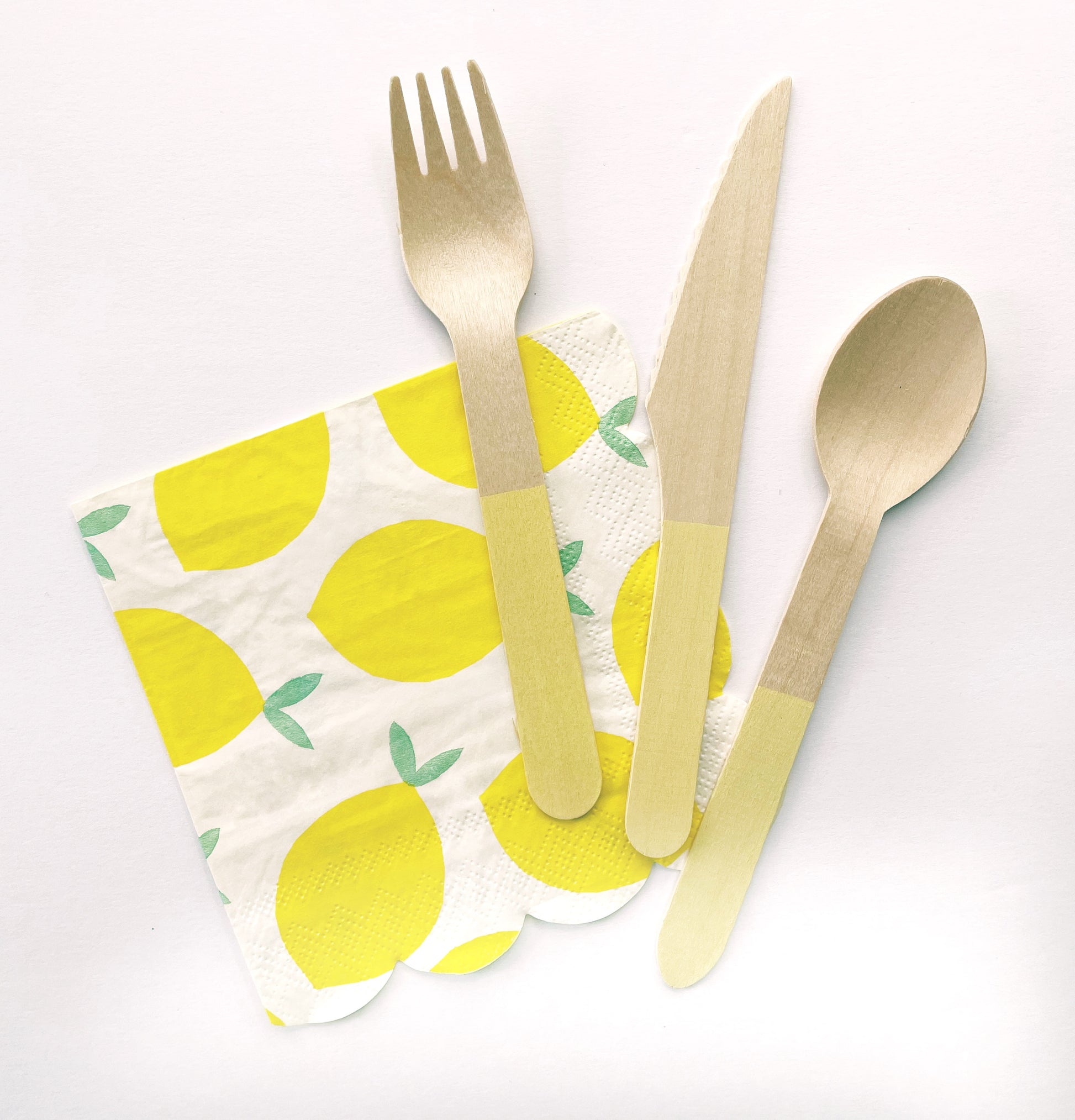 The Lemon Party Kit's napkins and utensils. The paper party napkins have a lemon pattern including yellow, green and white colours. The napkins have scalloped edges. The Lemon party kit's yellow dipped wooden utensils, including a fork, spoon and knife.