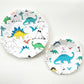 The large and small paper party plate dinosaur pattern features green, blue, yellow and red dinosaurs, volcanoes and palm trees. The party plates feature blue metallic elements, and are in the shape of a dodecagon.