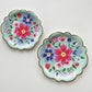 The Blossom Party Kit's paper party plates. The flower/floral pattern includes green, red and blue colours. The party plates have gold scalloped edges. This image shows the large and small plates.