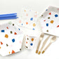 The complete Terrazzo Party Kit including large paper party plates, small paper party plates, paper cups, paper napkins, gold dipped wooden utensils and tall blue birthday candles. The terrazzo pattern includes blue, gold, orange and green colours. The party plates are in the shape of a hexagon.