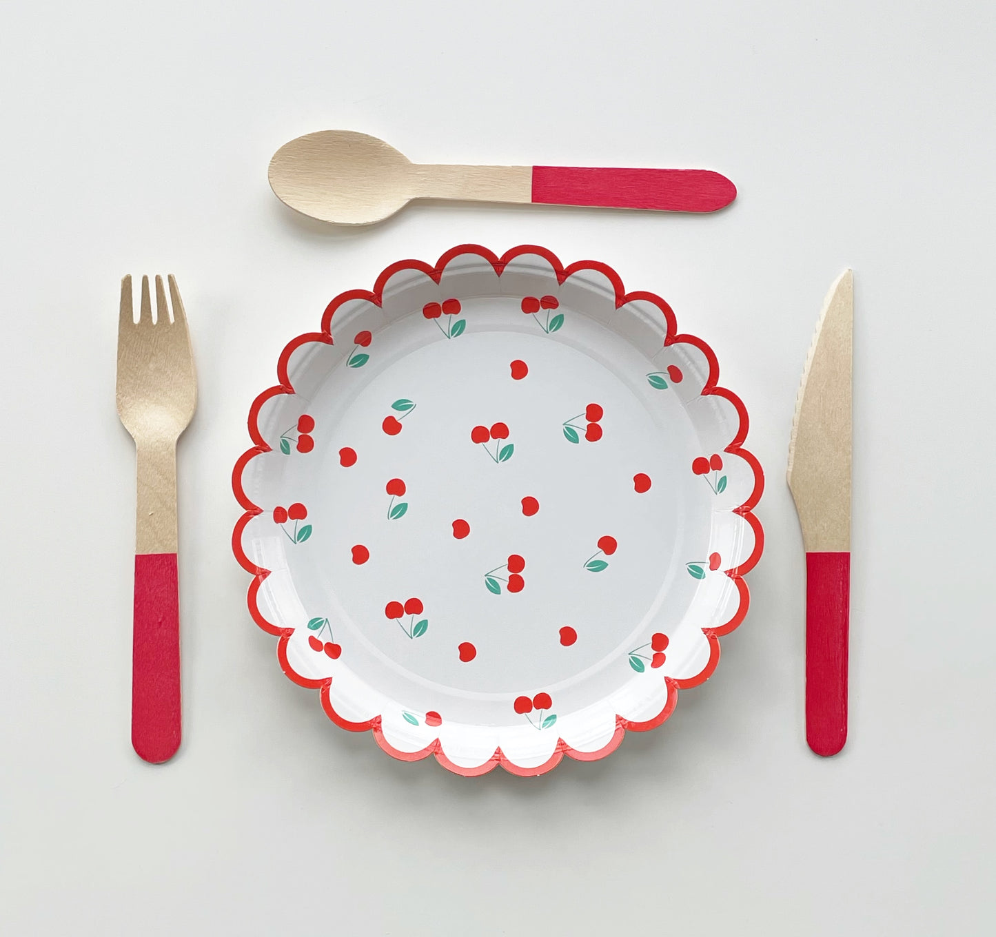 The small Cherry paper party plates have a red scalloped edge. The cherry pattern includes red, white and green colours.  Red dipped wooden utensils are arranged around the plate. This cutlery set includes knives, spoons and forks.