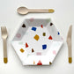 The small Terrazzo paper party plates are in the shape of a hexagon. The terrazzo pattern includes blue, gold, orange and green colours.  Gold dipped wooden utensils. This cutlery set includes knives, spoons and forks.