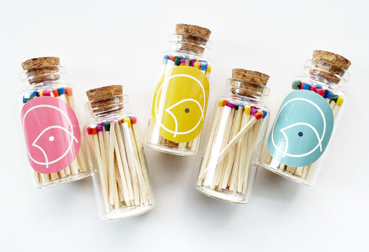 Five small glass jars filled with 30 rainbow colour tipped safety matches. The jars have a cork stopper and pink, yellow and blue stickers on them.