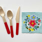 The Blossom Party Kit's napkins and utensils. The paper party napkins have a flower/floral pattern including green, red and blue colours. The napkins have scalloped edges. The Blossom party kit's red dipped wooden utensils, including a fork, spoon and knife.