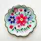 The Blossom Party Kit's paper party plates. The flower/floral pattern includes green, red and blue colours. The party plates have gold scalloped edges.