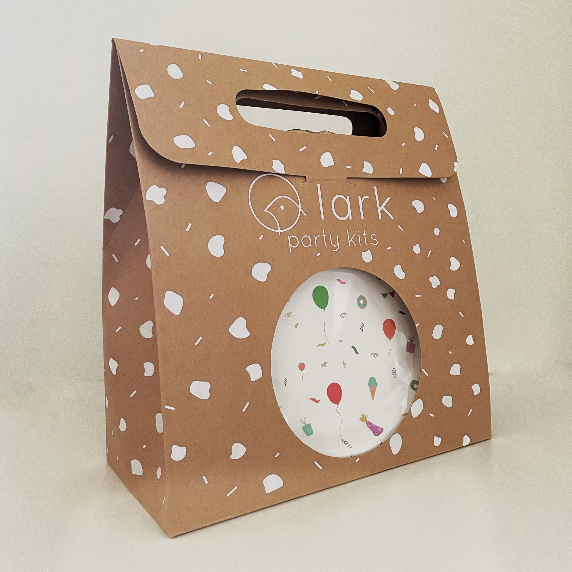 The Celebration Party Kit in its kraft brown and white carry box. The box features a handle and a circular opening where you can see the contents inside.