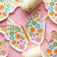 Parts of the Butterfly Party Kit including large pink and gold paper party plates, small butterfly shaped paper party plates and paper cups. The Butterfly pattern includes pink, gold, blue, green and orange colours. The large party plates are in the shape of an octagon.