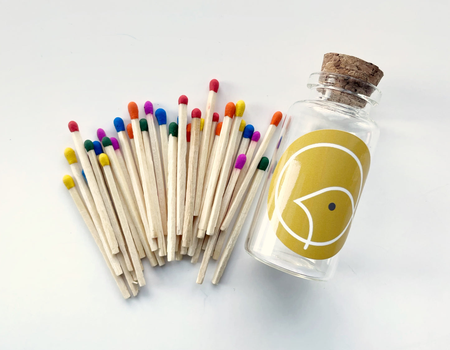 One small glass jar with 30 rainbow colour tipped safety matches laid out beside it. The jar has a cork stopper and a yellow label on it.