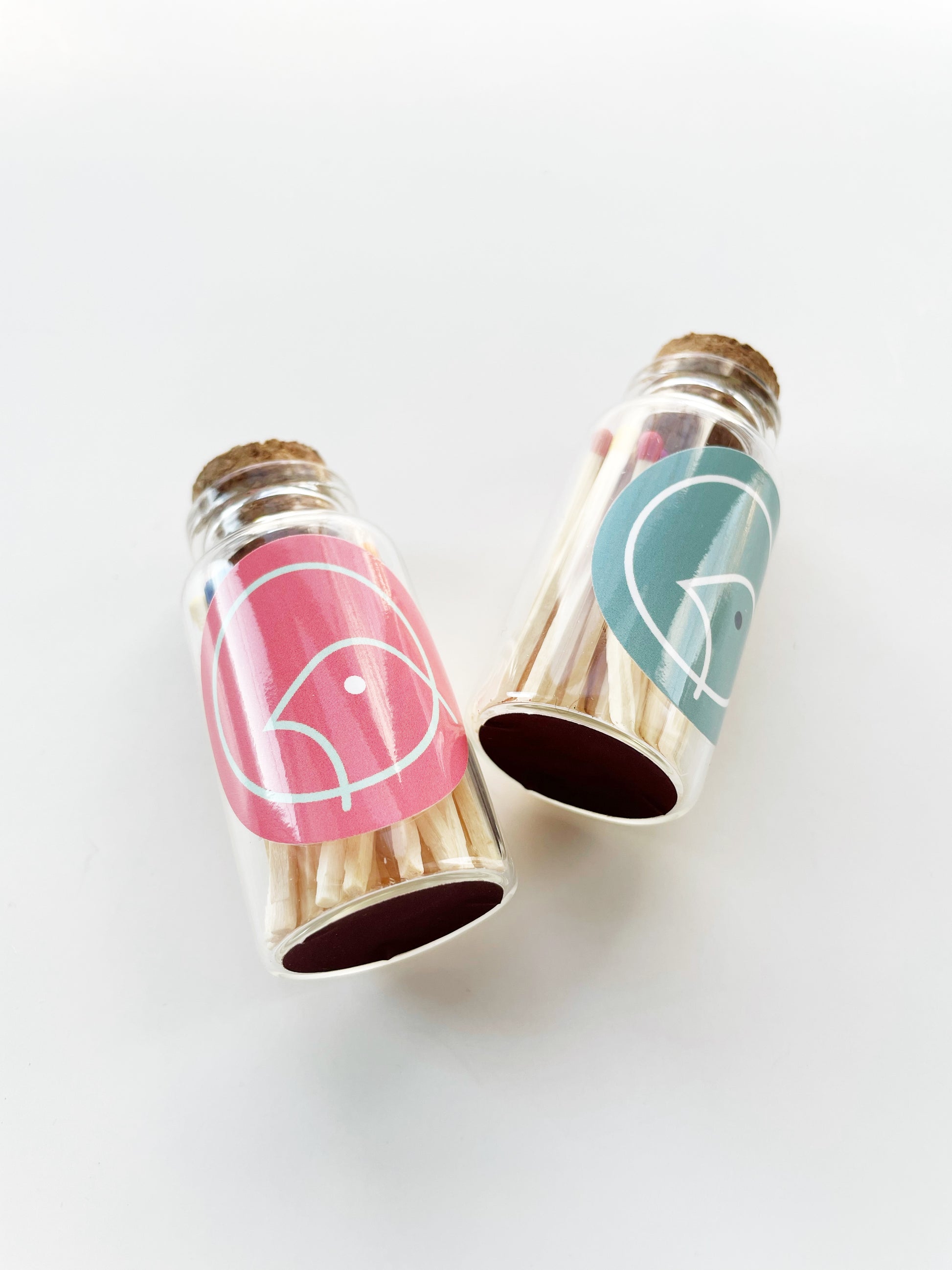 Two small glass jars with 30 rainbow colour tipped safety matches in them. The jars have a cork stopper and have blue and pink labels on them. The jars have strike pads on their base.