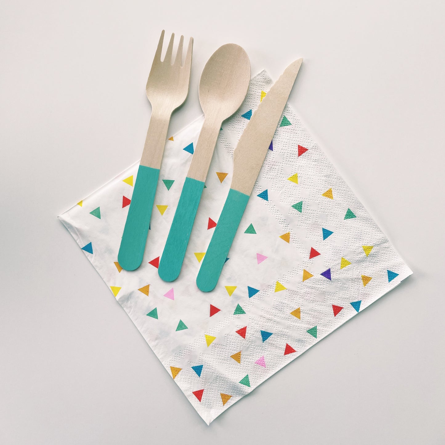 Paper party napkins with a triangle pattern featuring green, orange, pink, blue and yellow colours.  Teal dipped wooden utensils.