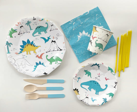 The complete Dinosaur Party Kit including large paper party plates, small paper party plates, paper cups, blue and white paper napkins, blue dipped wooden utensils and tall yellow birthday candles. The dinosaur pattern features green, blue, yellow and red dinosaurs, volcanoes and palm trees. The party plates feature blue metallic elements, and are in the shape of a dodecagon.  