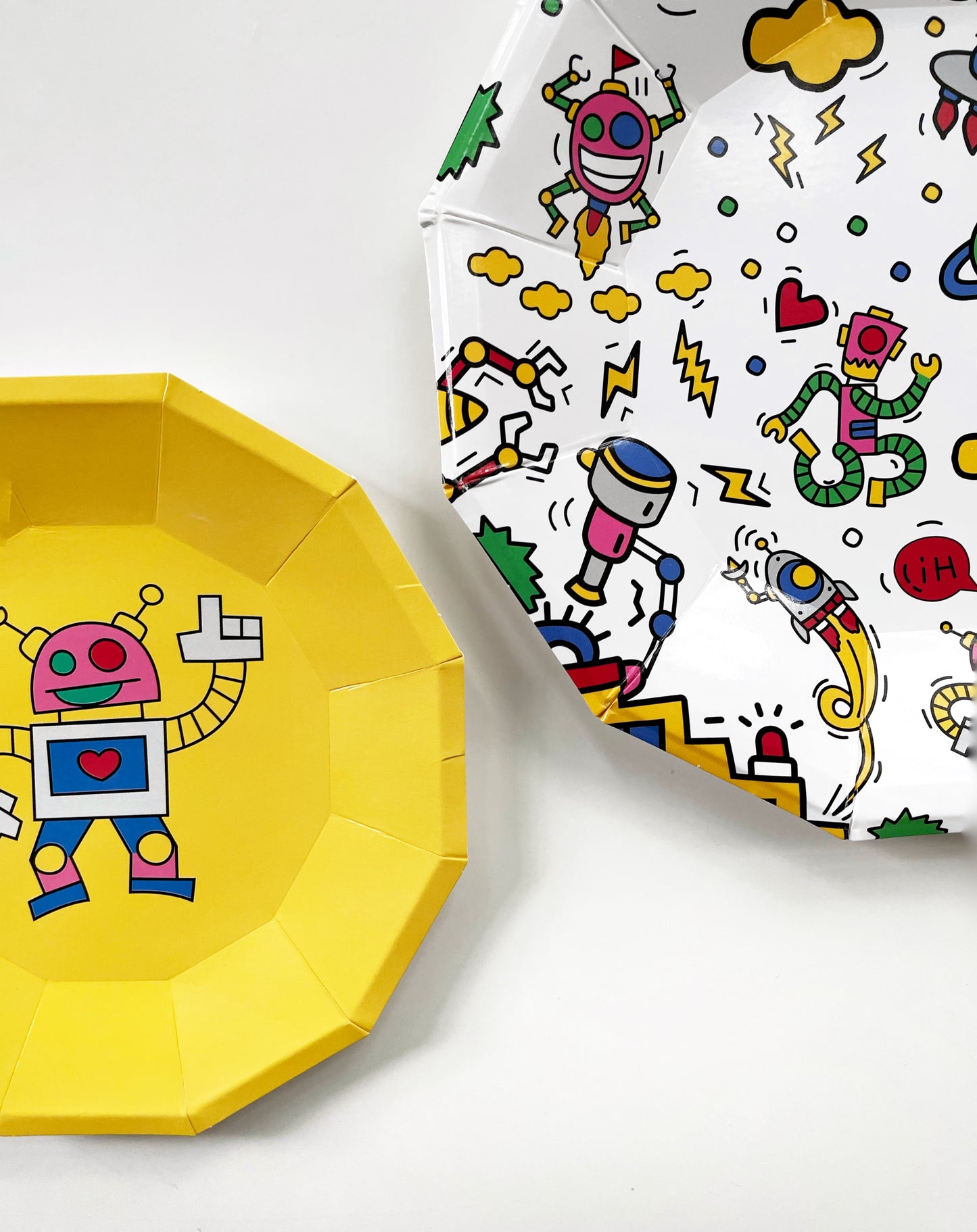 Close up of the large and small robot paper party plates. They are in the shape of a dodecagon. The robot pattern includes planets, robots, spaceships and clouds in green, blue, red, yellow and pink colours.  