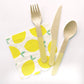 The Lemon Party Kit's napkins and utensils. The paper party napkins have a lemon pattern including yellow, green and white colours. The napkins have scalloped edges. The Lemon party kit's yellow dipped wooden utensils, including a fork, spoon and knife.