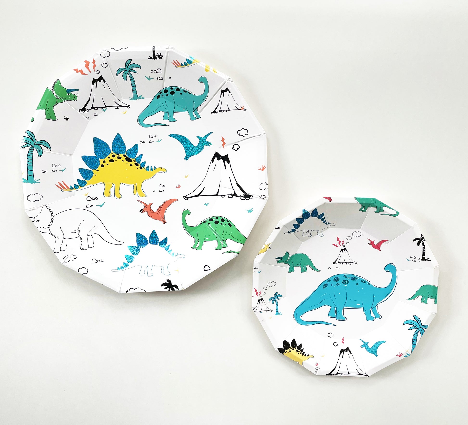 The large and small paper party plate dinosaur pattern features green, blue, yellow and red dinosaurs, volcanoes and palm trees. The party plates feature blue metallic elements, and are in the shape of a dodecagon.