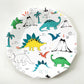 The large paper party plate dinosaur pattern features green, blue, yellow and red dinosaurs, volcanoes and palm trees. The party plates feature blue metallic elements, and are in the shape of a dodecagon.