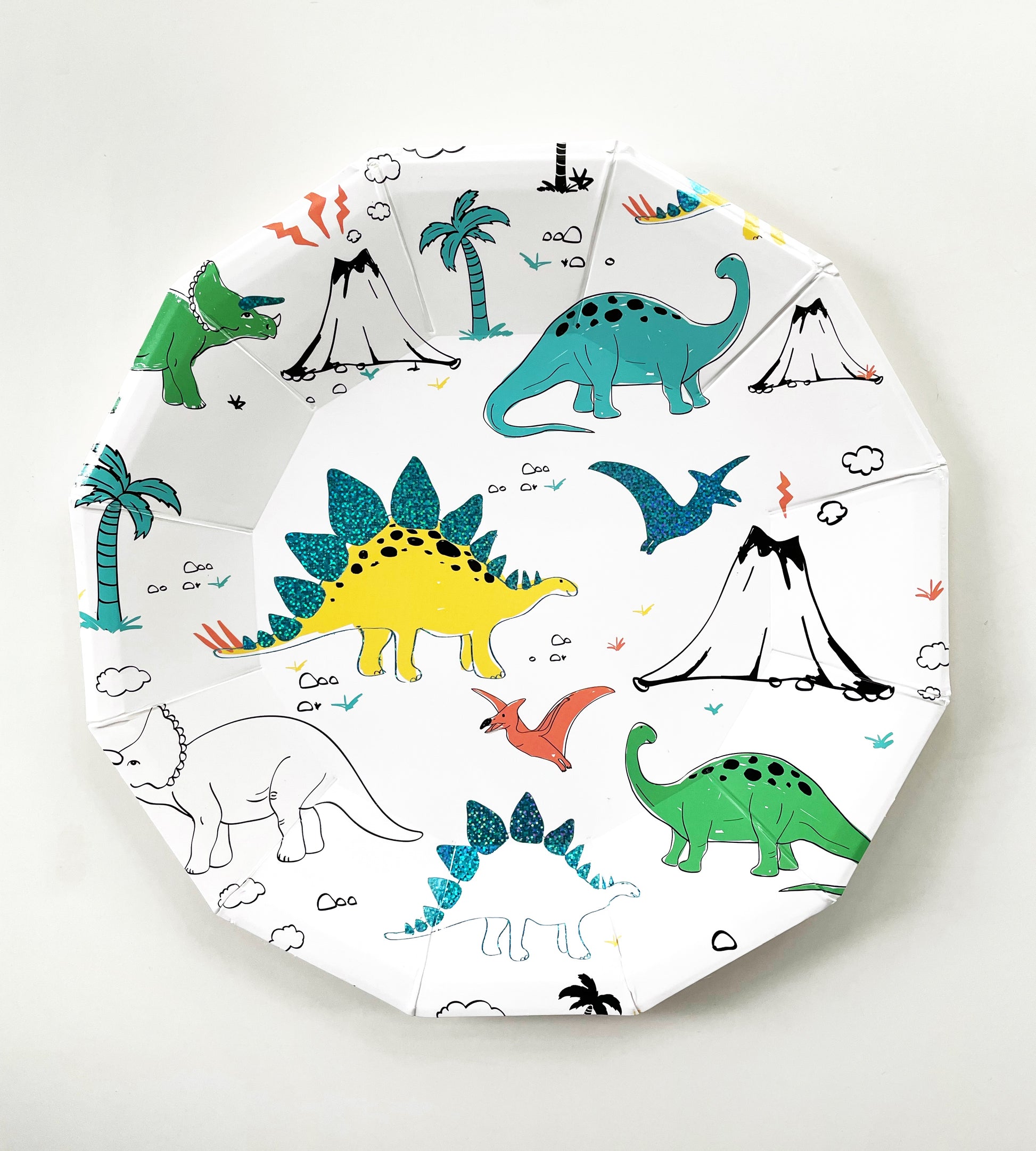The large paper party plate dinosaur pattern features green, blue, yellow and red dinosaurs, volcanoes and palm trees. The party plates feature blue metallic elements, and are in the shape of a dodecagon.