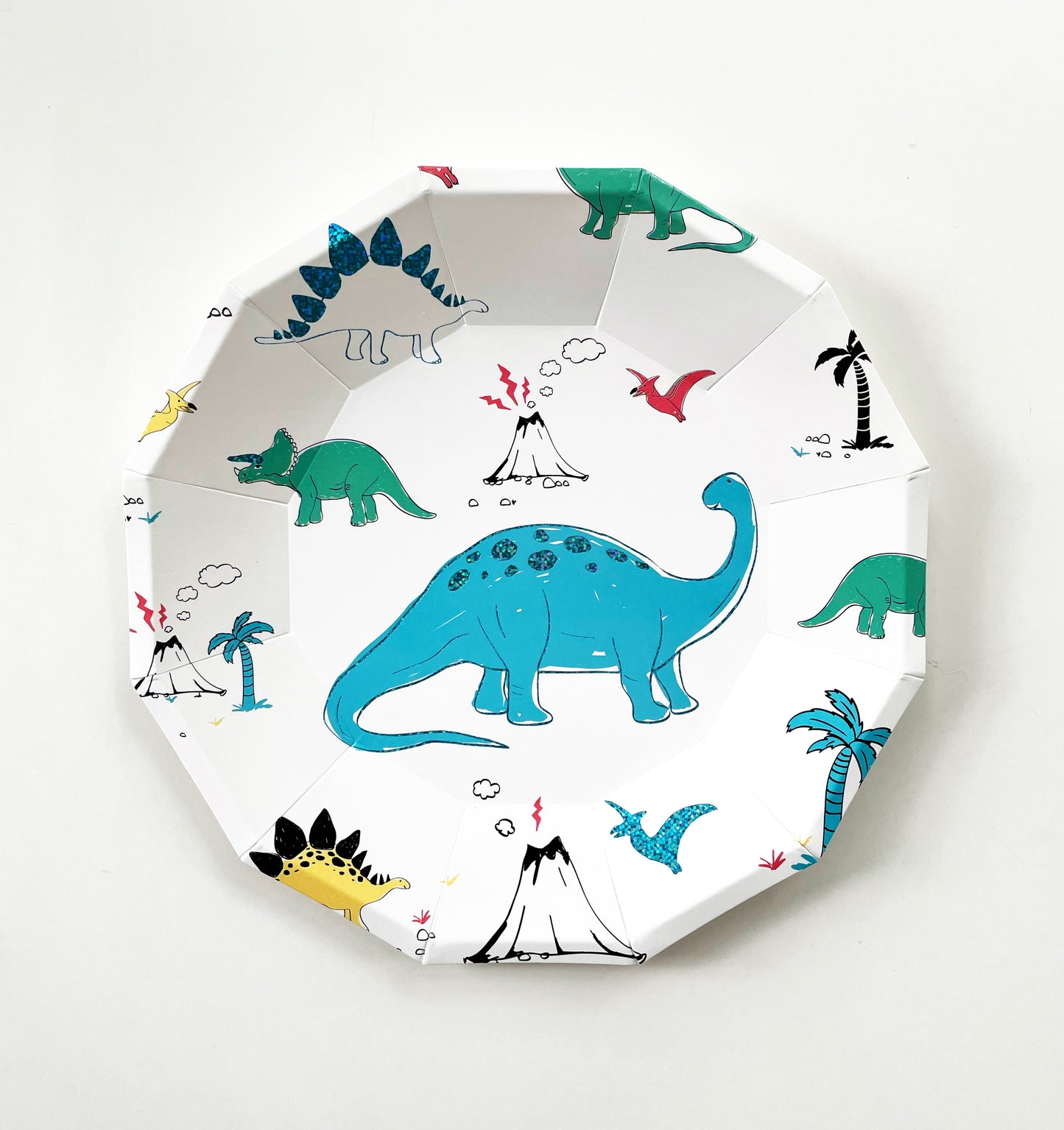 The small paper party plate dinosaur pattern features green, blue, yellow and red dinosaurs, volcanoes and palm trees. The party plates feature blue metallic elements, and are in the shape of a dodecagon.